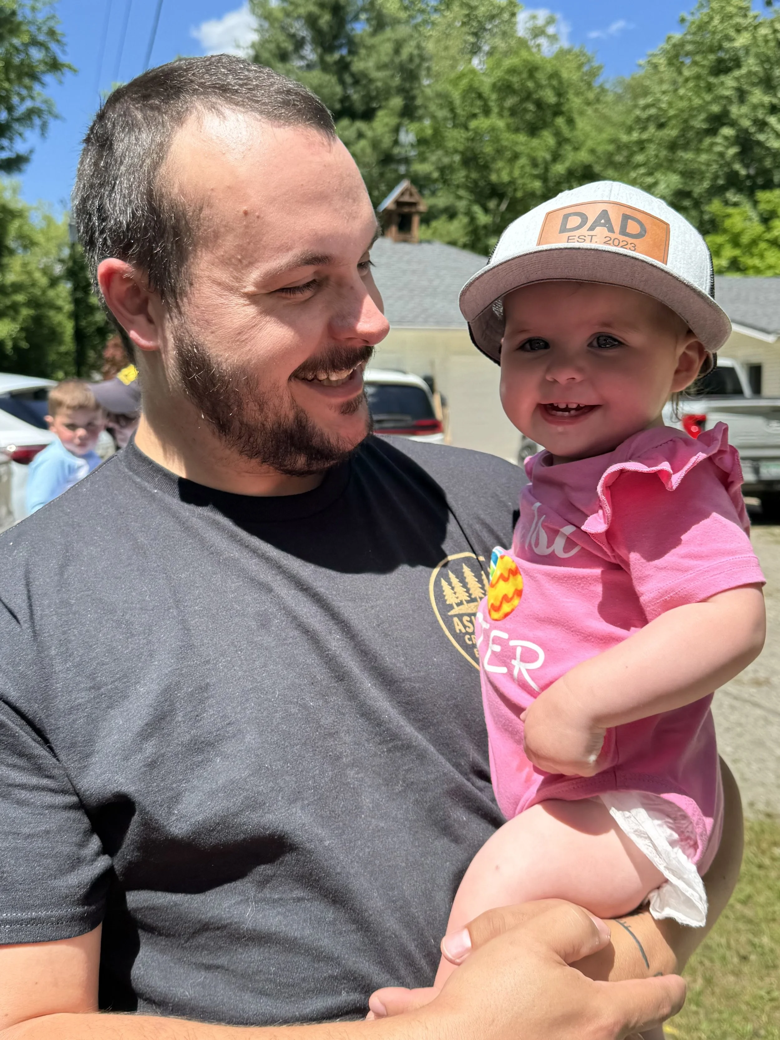 A man holding a smiling toddler girl outdoors during daytime with green trees, a house, cars, and a boy in the background.