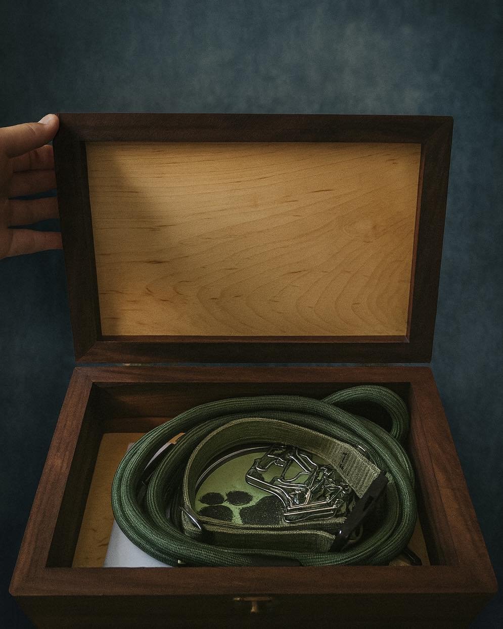 A wooden box opened to reveal a green fabric strap and a metal clamp inside.