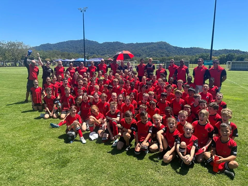 A large group of children and coaches in red and black soccer uniforms posing for a team photo on a sunny day at a soccer field, with mountains and a blue sky in the background.