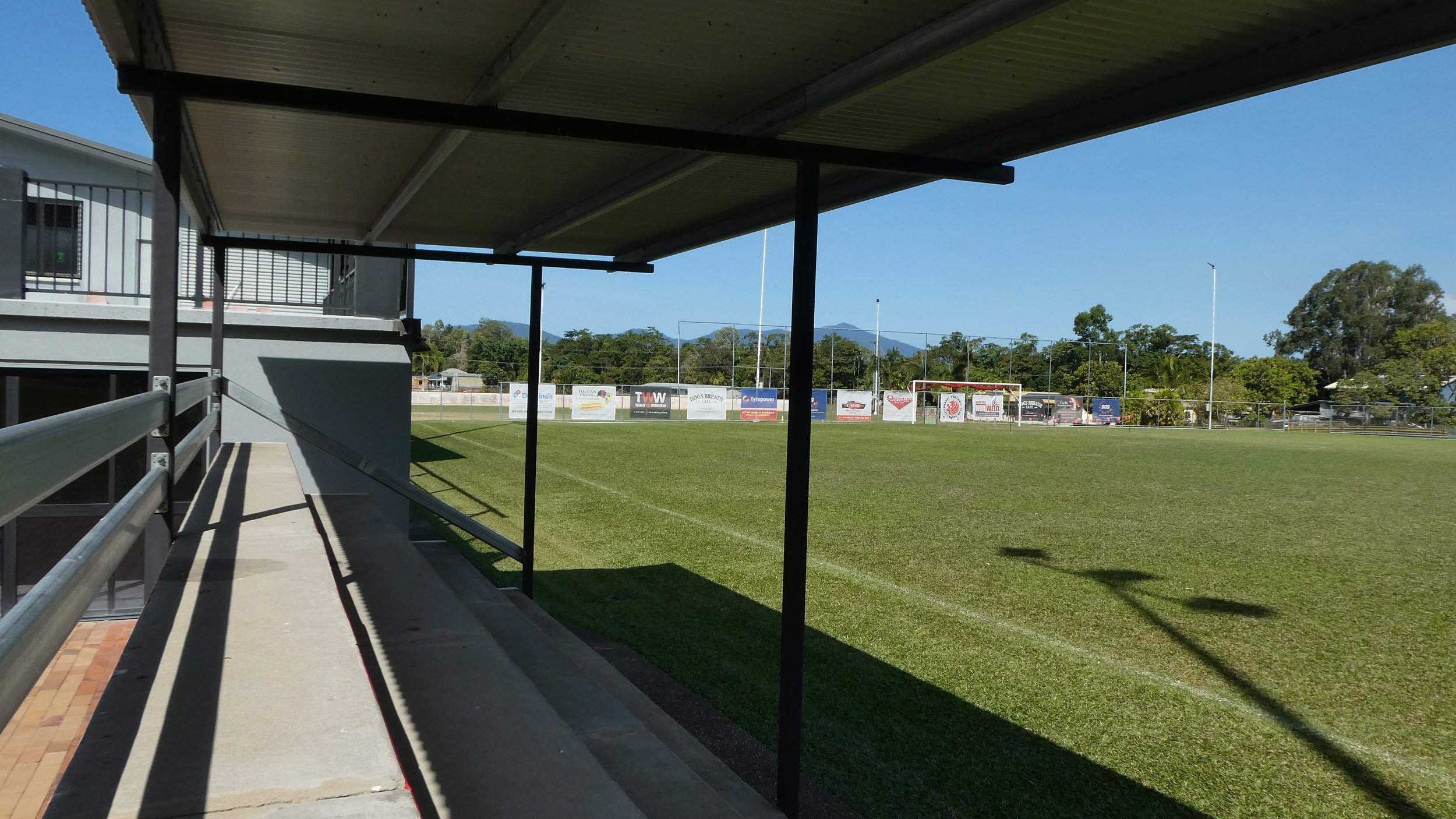 View of a sports field from a shaded spectator area with a building on the left and advertisements on the far fence with trees and mountains in the background.