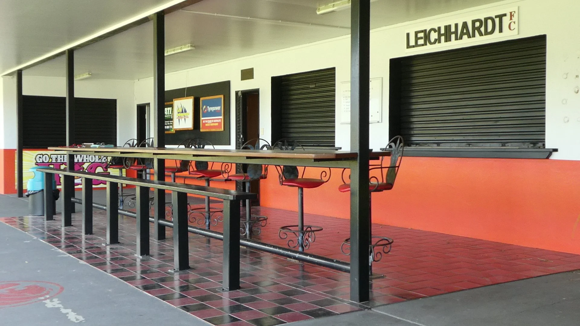 Empty outdoor seating area with a counter, chairs with red cushions, and black metal railings outside closed shops with black roller shutters.