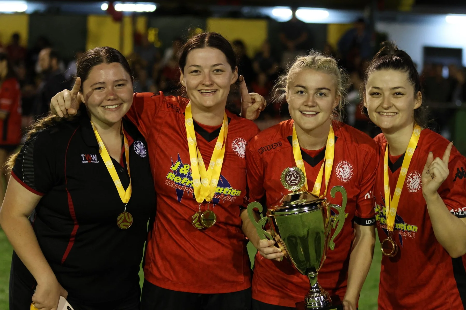Four female soccer players in red and black uniforms celebrate with medals and a trophy on a field at night.