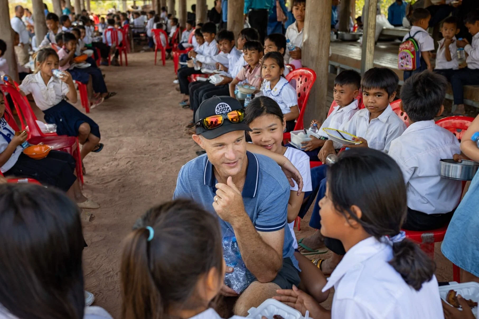 A diverse group of school children sitting on red plastic chairs and eating lunch, with a man in a blue shirt and cap engaging with them in an outdoor setting with wooden posts and dirt ground.