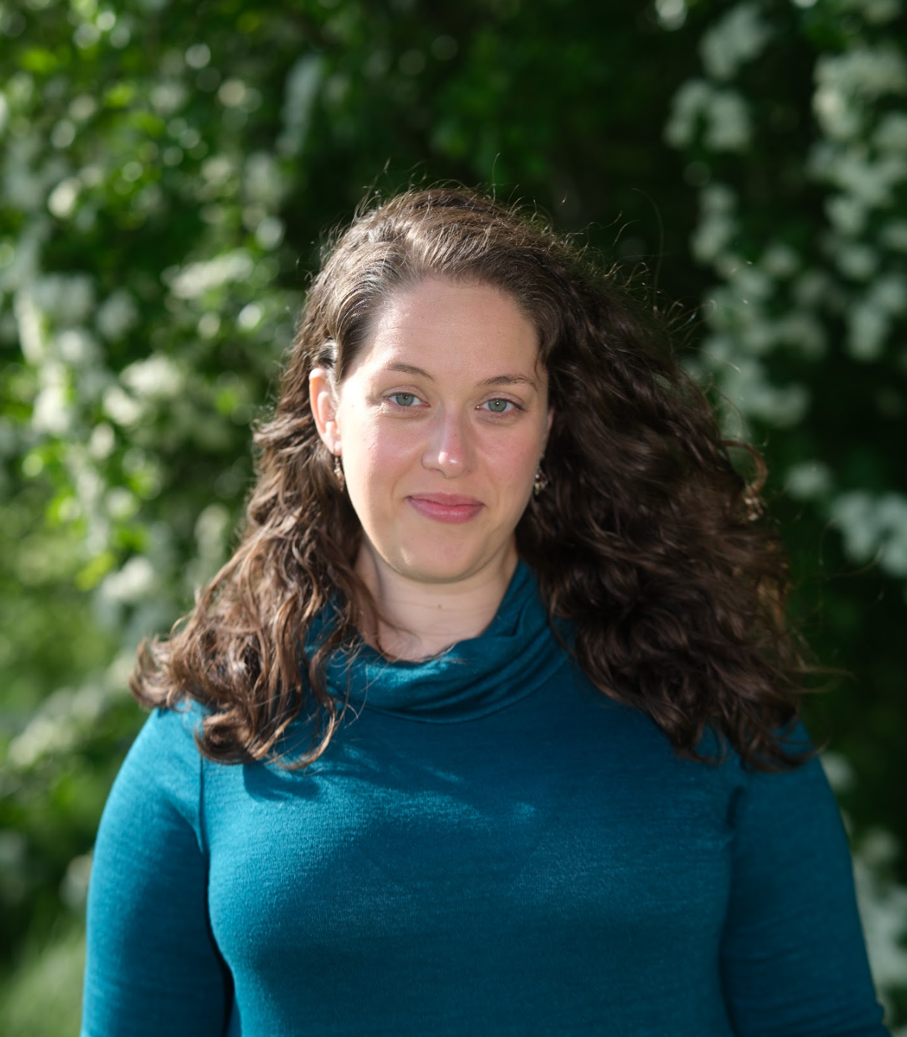 Image of a woman smiling with curly brown hair, wearing a teal knit top. She's looking directly at the camera, warmly. The background is blurred green leaves of a tree.