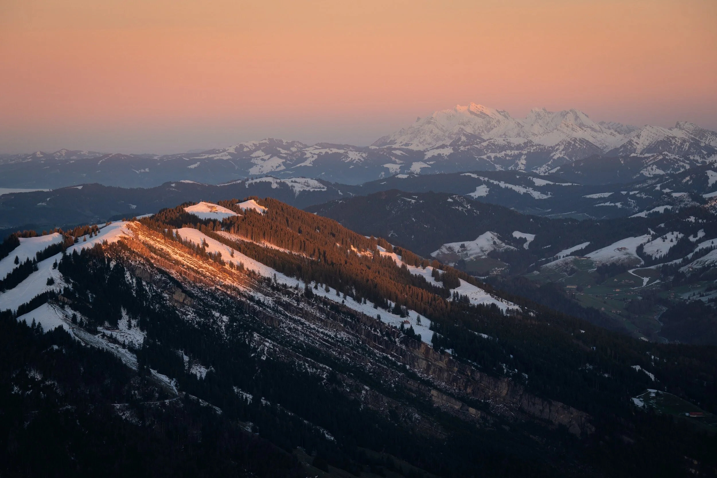 Sunset over snow-covered mountains and hills with a pink sky.