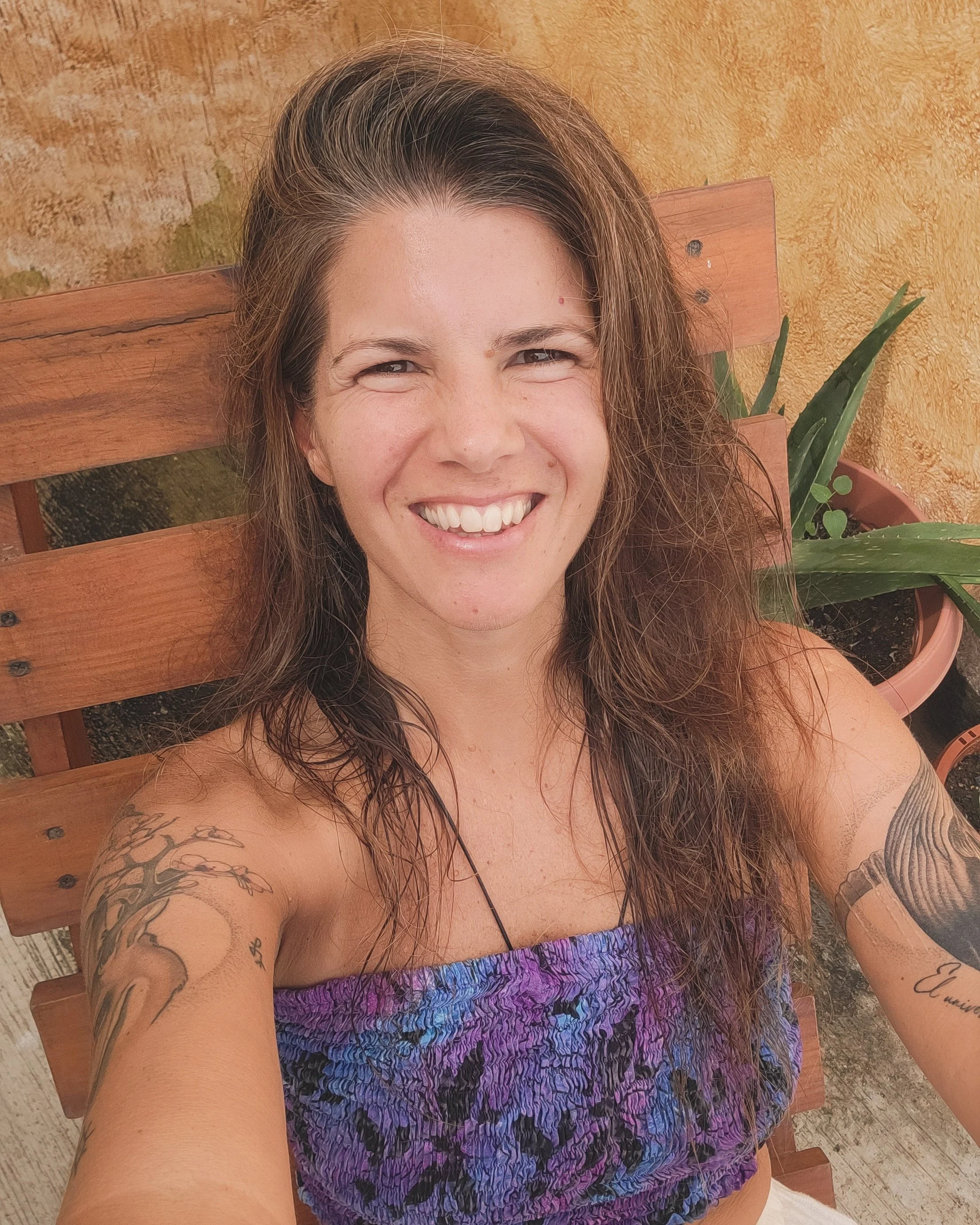 A smiling woman with long brown hair and tattoos on her arms, wearing a strapless purple and blue patterned top, taking a selfie outdoors while sitting on a wooden bench with potted plants nearby.