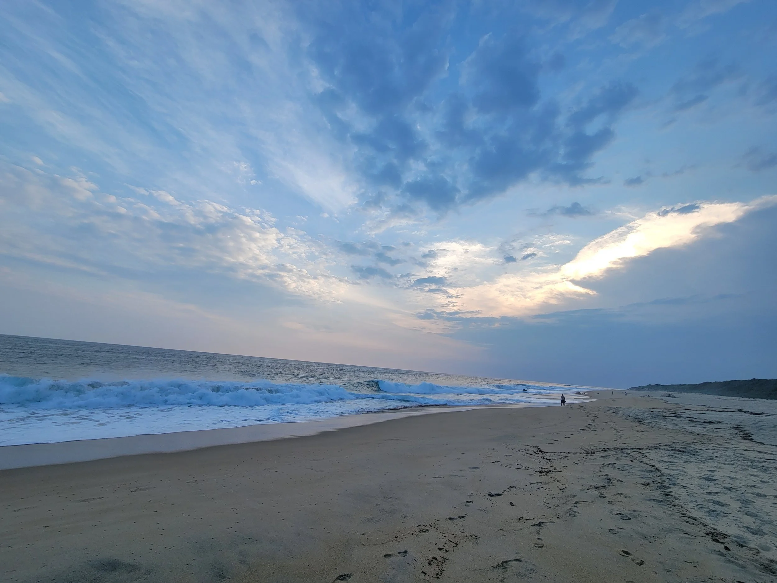 A peaceful beach scene with sand, ocean waves, and a partly cloudy sky during sunset or sunrise.