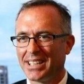 A man with glasses and short hair smiling in front of a cityscape background.