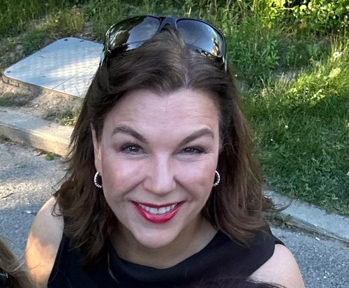 Close-up of a woman smiling, wearing sunglasses on her head, with dark brown hair, red lipstick, earrings, and a black top, outdoors on a sunny day.