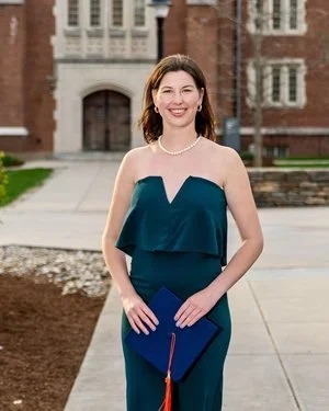 A young woman in a teal strapless dress holding a diploma, standing outside in front of a brick building, celebrating graduation.