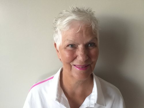 A smiling elderly woman with short white hair, wearing a white shirt with pink stripes on the shoulders, standing against a plain light-colored wall.