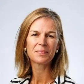 A middle-aged woman with blonde hair and a neutral expression, wearing a black and white striped top and pearl earrings, against a plain white background.