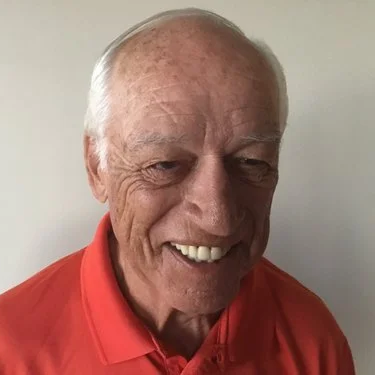Close-up of smiling elderly man with white hair wearing a red polo shirt against a plain background.