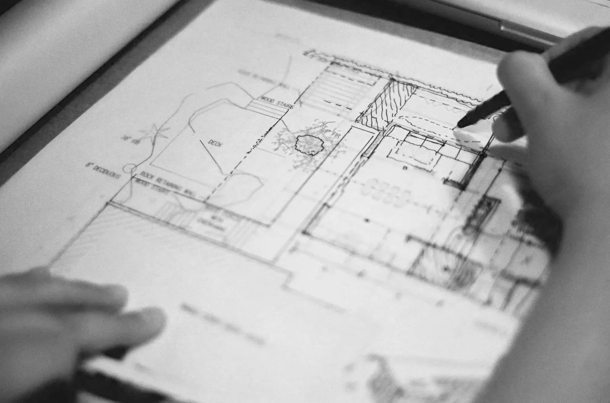 A technical drawing or blueprint of a house layout on paper with a person’s hand holding a pen, appearing to make notes or adjustments.