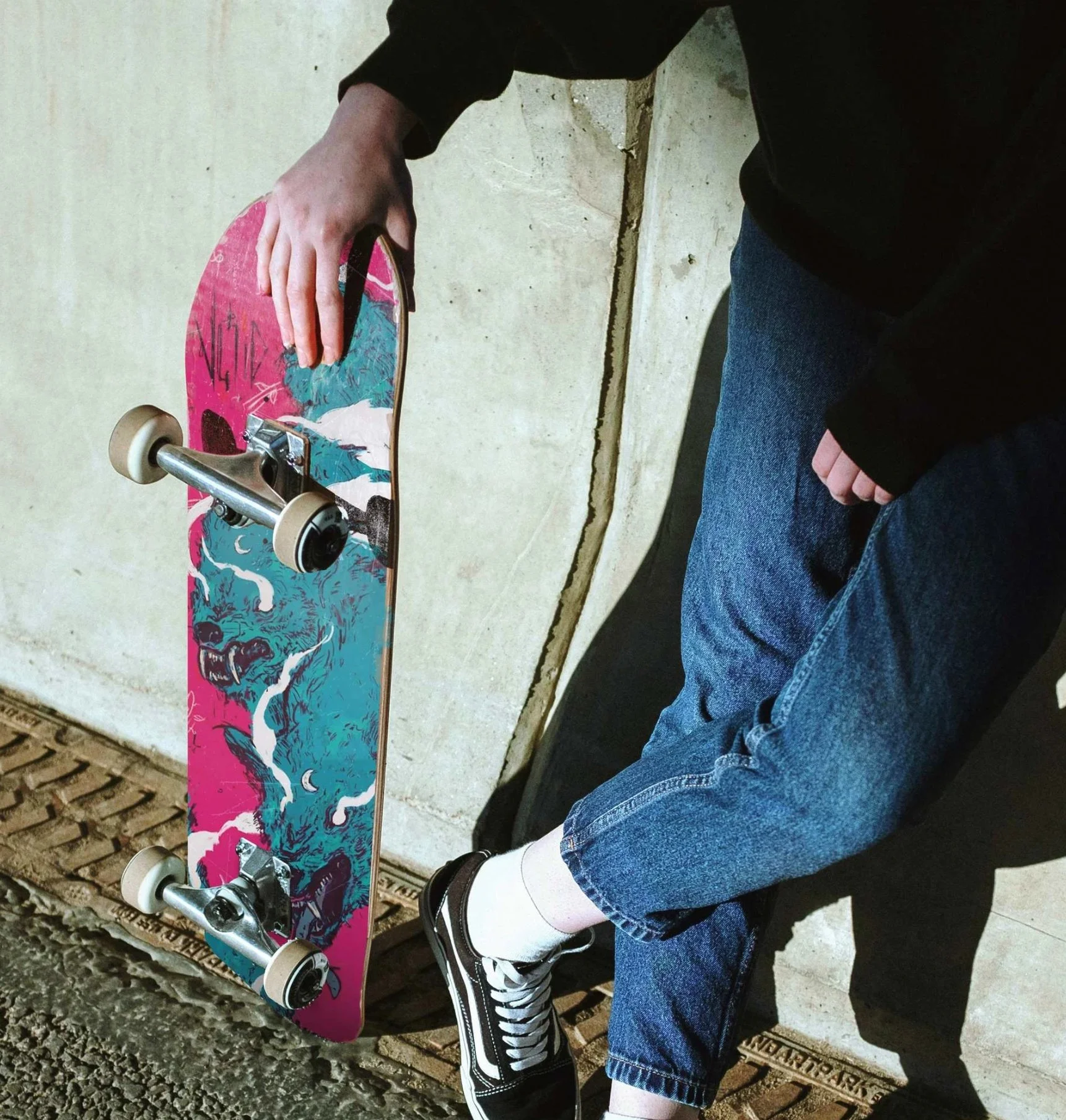 Person holding a skateboard with a pink and blue graphic design, standing against a concrete wall.