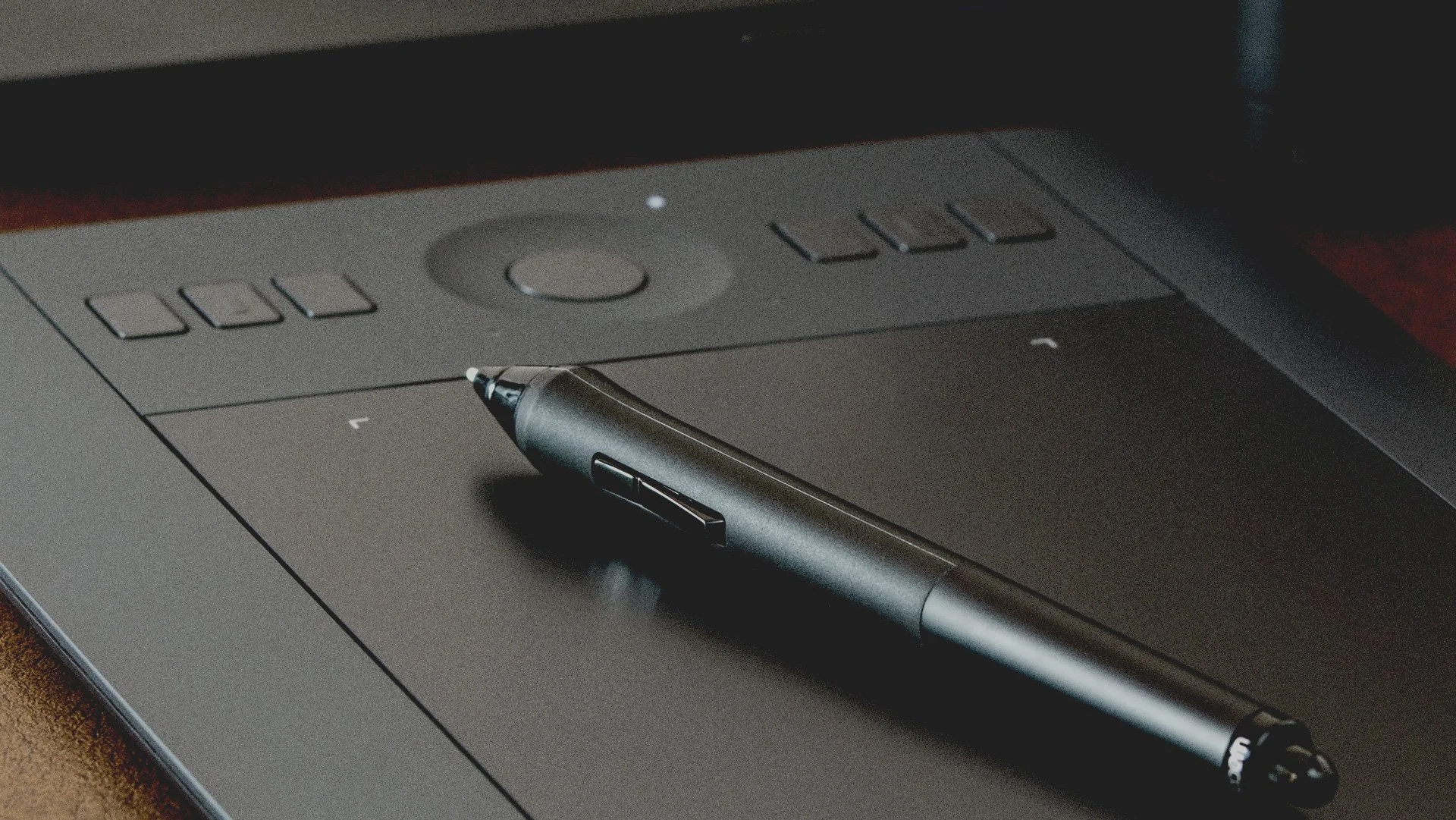 A black digital pen resting on a closed black drawing tablet with buttons and a circular control, on a wooden surface.