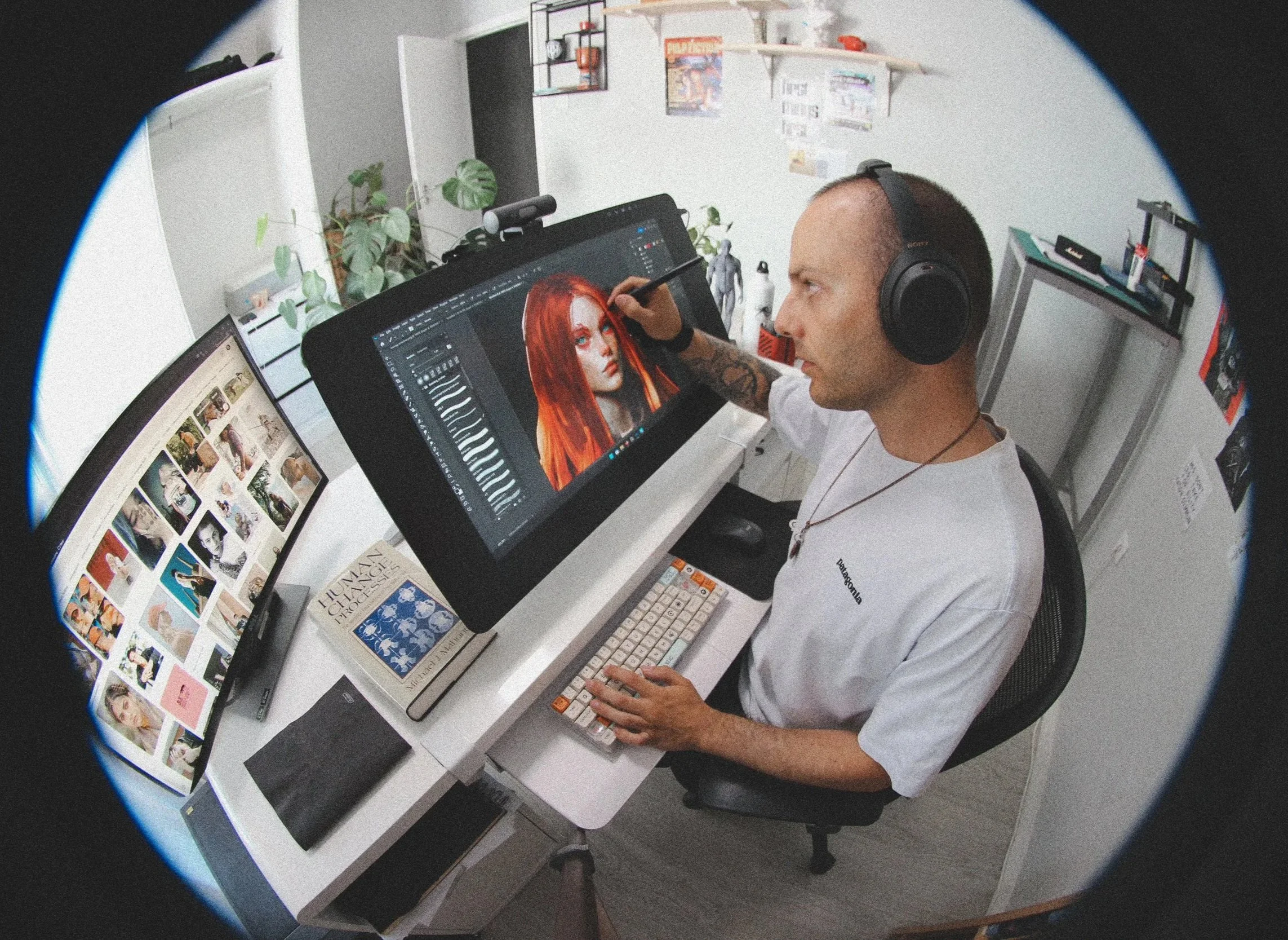 A person with a tattoo on their arm and wearing headphones is working on a digital portrait of a woman with red hair on a graphics tablet. The workspace includes a computer monitor displaying a grid of images, potted plants, and cluttered shelves in the background.