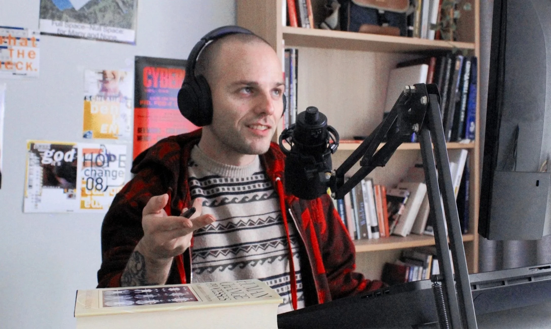 A man with a shaved head and tattoos on his arm is wearing large headphones and speaking into a microphone in a room with bookshelves. There are posters and magazines on the wall behind him.