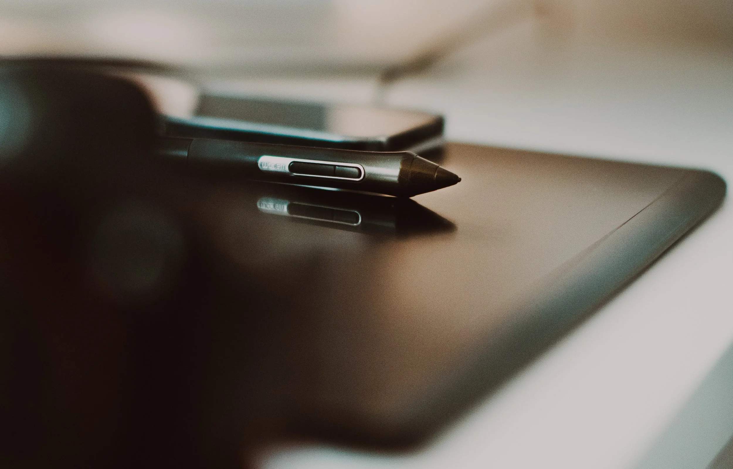 Close-up of a stylus pen resting on a digital tablet with a smartphone and other objects in the blurred background.