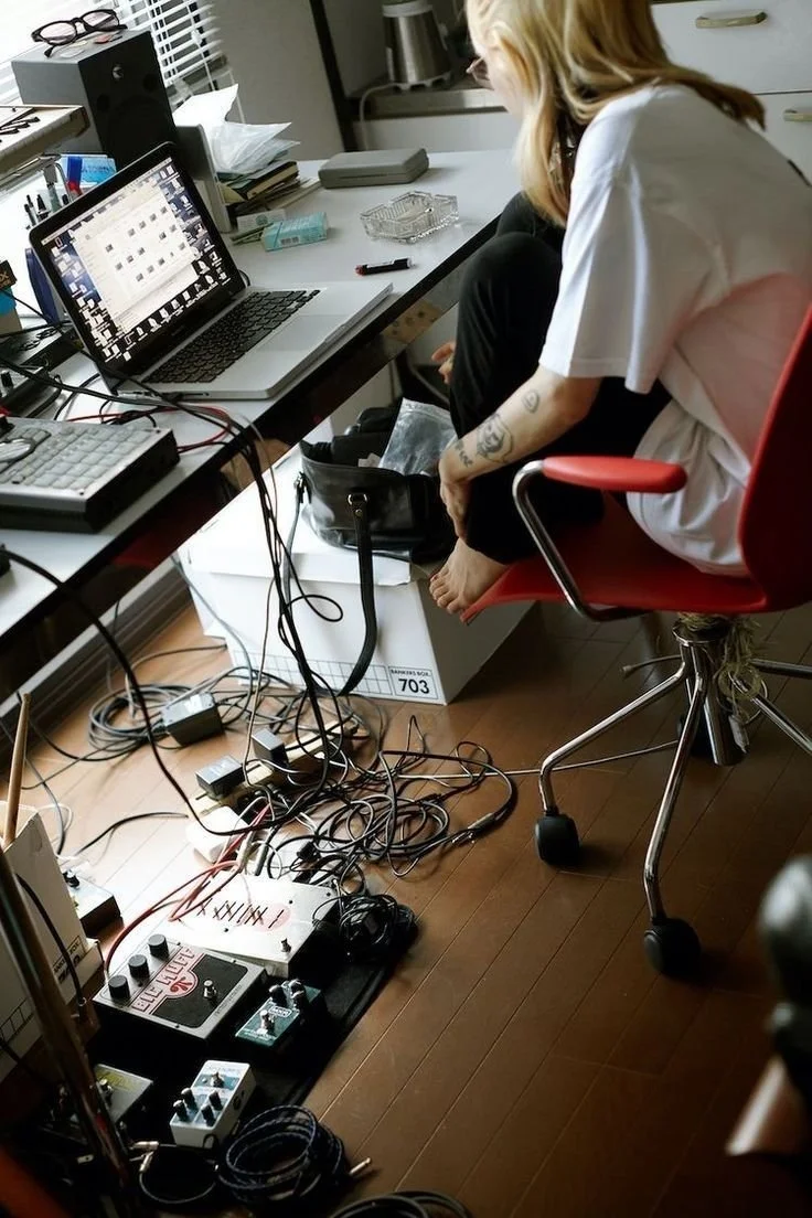 A person seated at a cluttered desk with electronic equipment, cables, and a laptop, with tattoos visible on their arm.