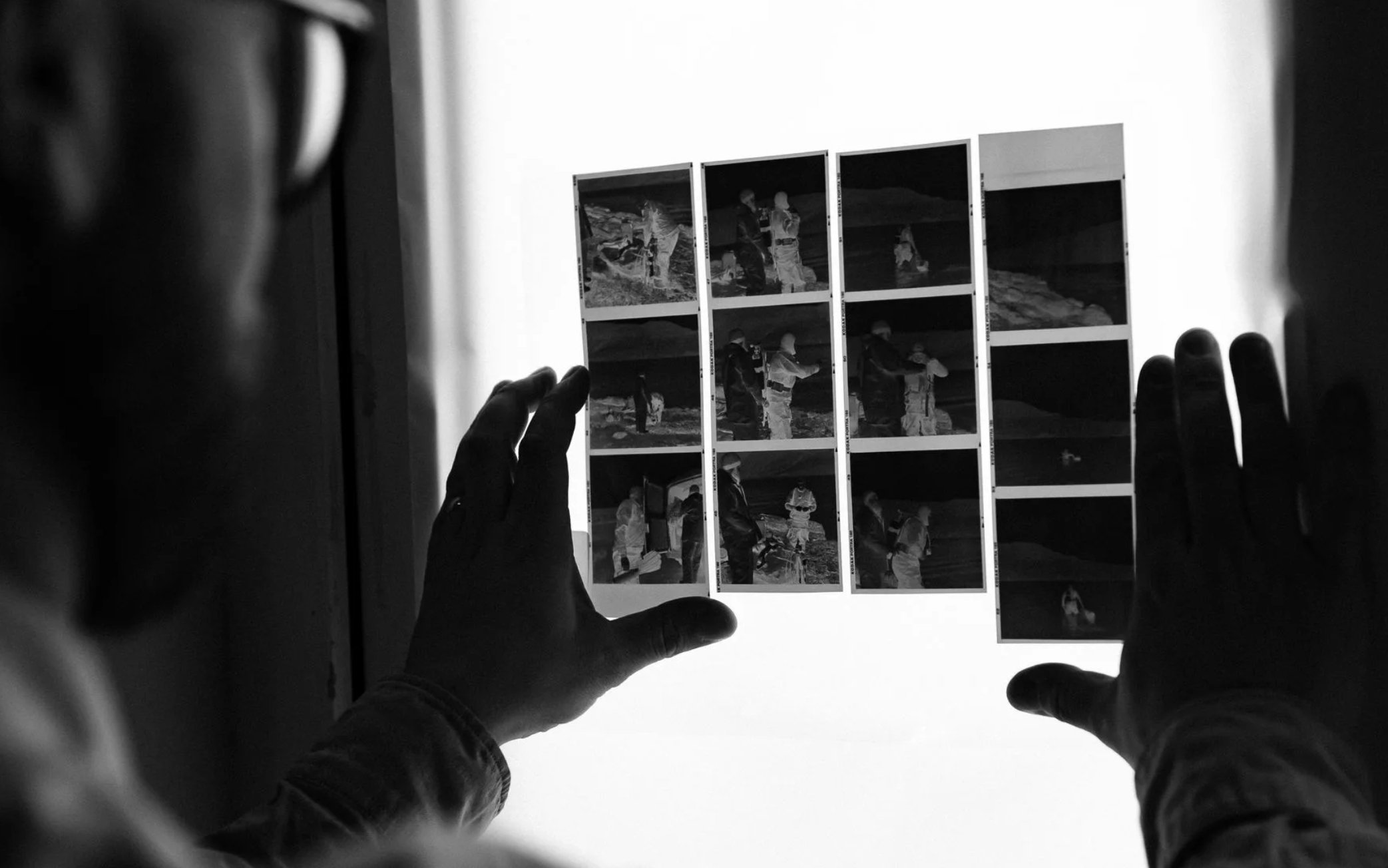 Black and white photo of individuals reviewing contact sheets with multiple film photographs, viewed through a backlit lightbox.