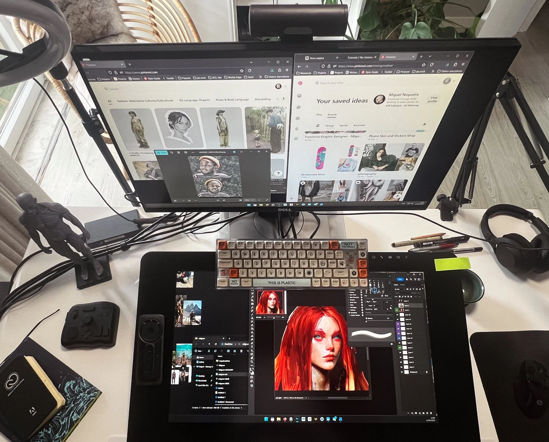 A workspace with a dual-monitor setup, a keyboard, a drawing tablet showing a digital portrait of a red-haired woman, and various art supplies on a white desk. There are two figurines on the left side of the desk and a window with a view outside.