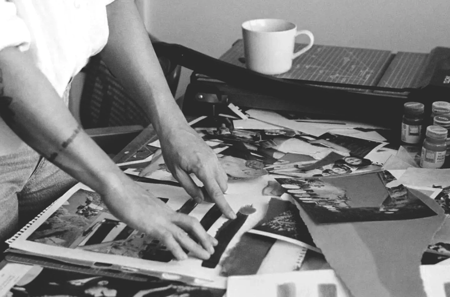 A person sorting through a collection of photographs and paper memorabilia on a cluttered desk in black and white.