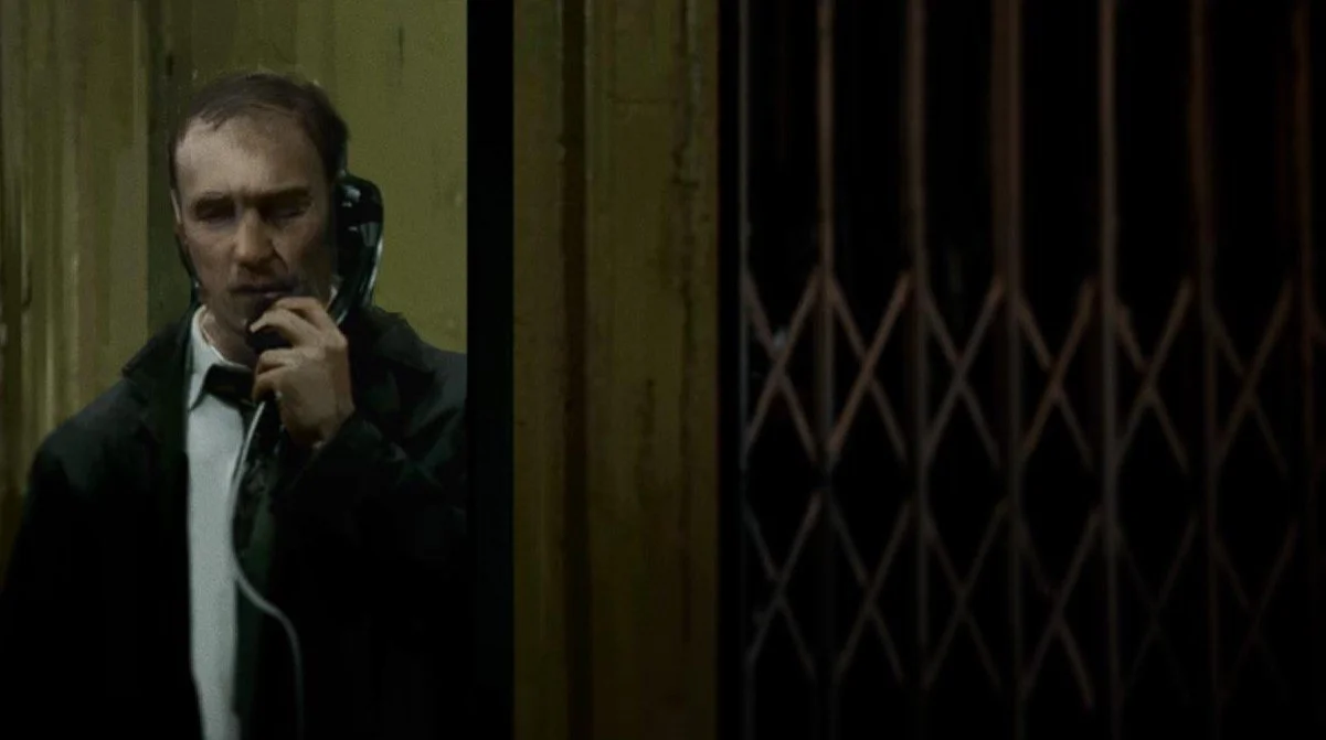 A man in formal attire using a vintage wall-mounted telephone in an elevator or lift with wooden paneling.
