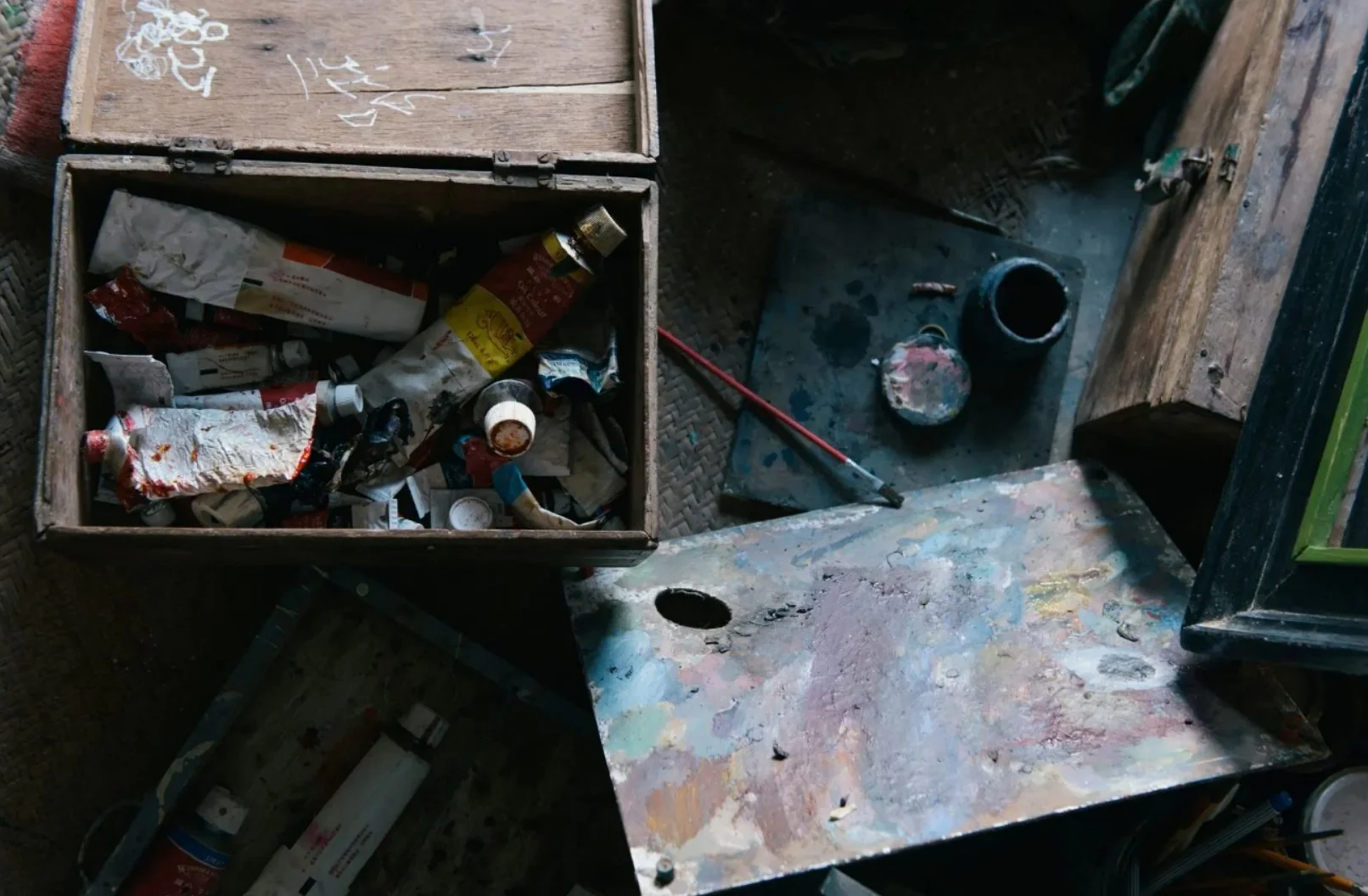 An art workspace with a paint-splattered table, a small open wooden box filled with used paint tubes and supplies, and various art tools and containers around.