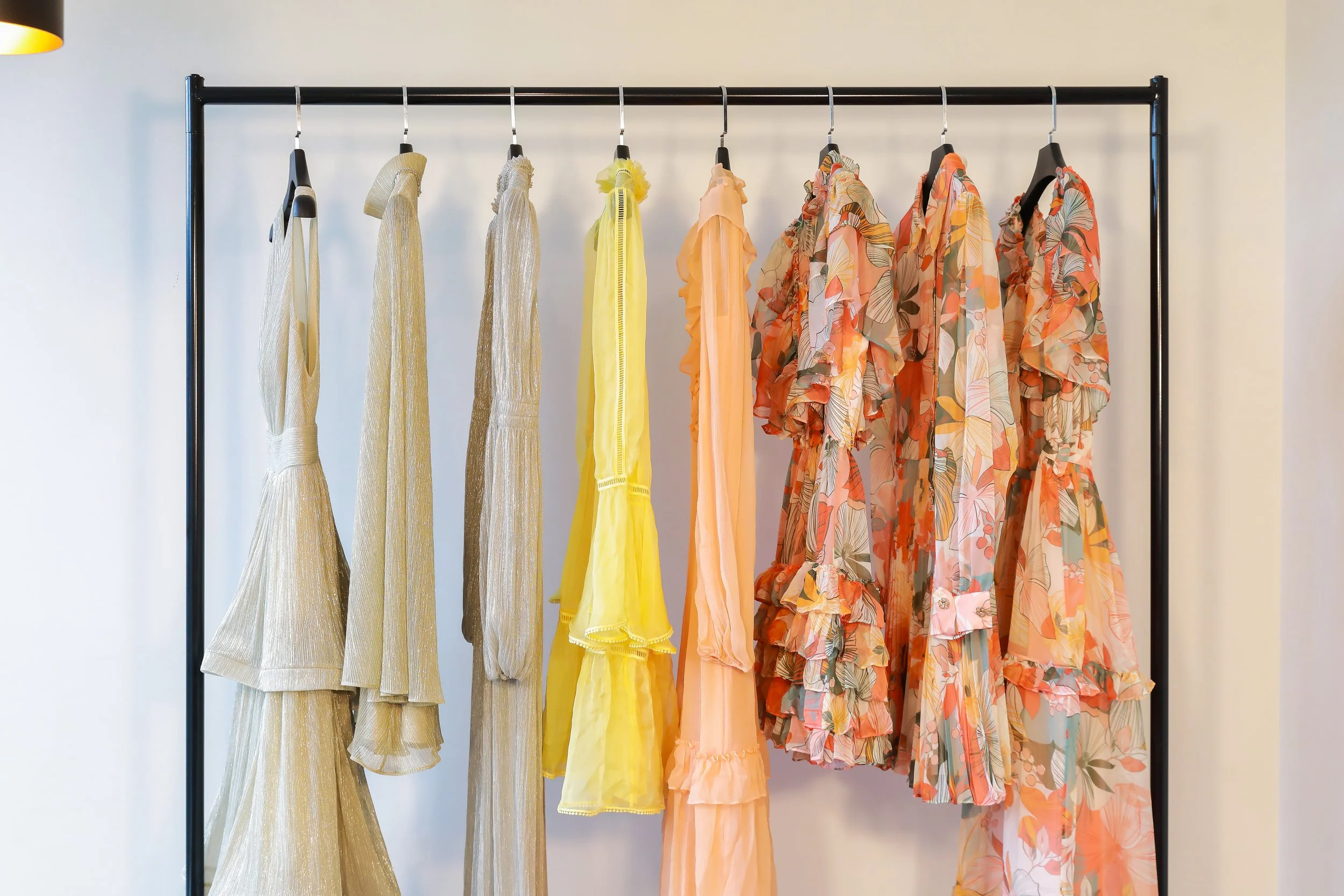 Colorful dresses hanging on a black clothing rack, ranging from gold to pastel yellow, orange, and floral prints in peach, coral, and pink tones.