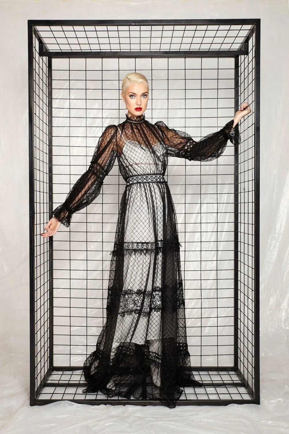 A woman with platinum blonde hair and red lipstick standing inside a black wireframe cage, wearing a black lace dress with sheer long sleeves and a white underdress.