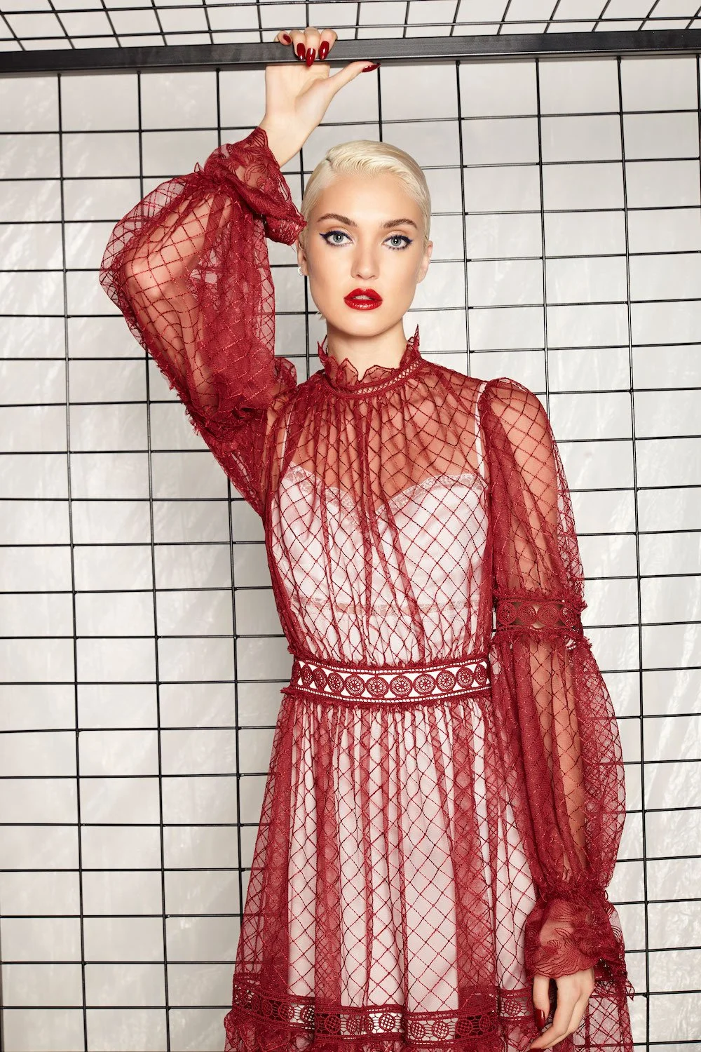 A woman with platinum blonde hair and red lipstick wearing a sheer red lace dress with long sleeves, standing against a white grid wall.