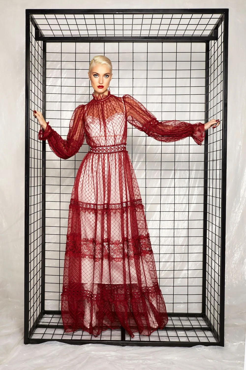 A woman with platinum blonde hair and red lipstick wearing a red lace dress with sheer long sleeves, standing inside a black metal cube frame against a white background.