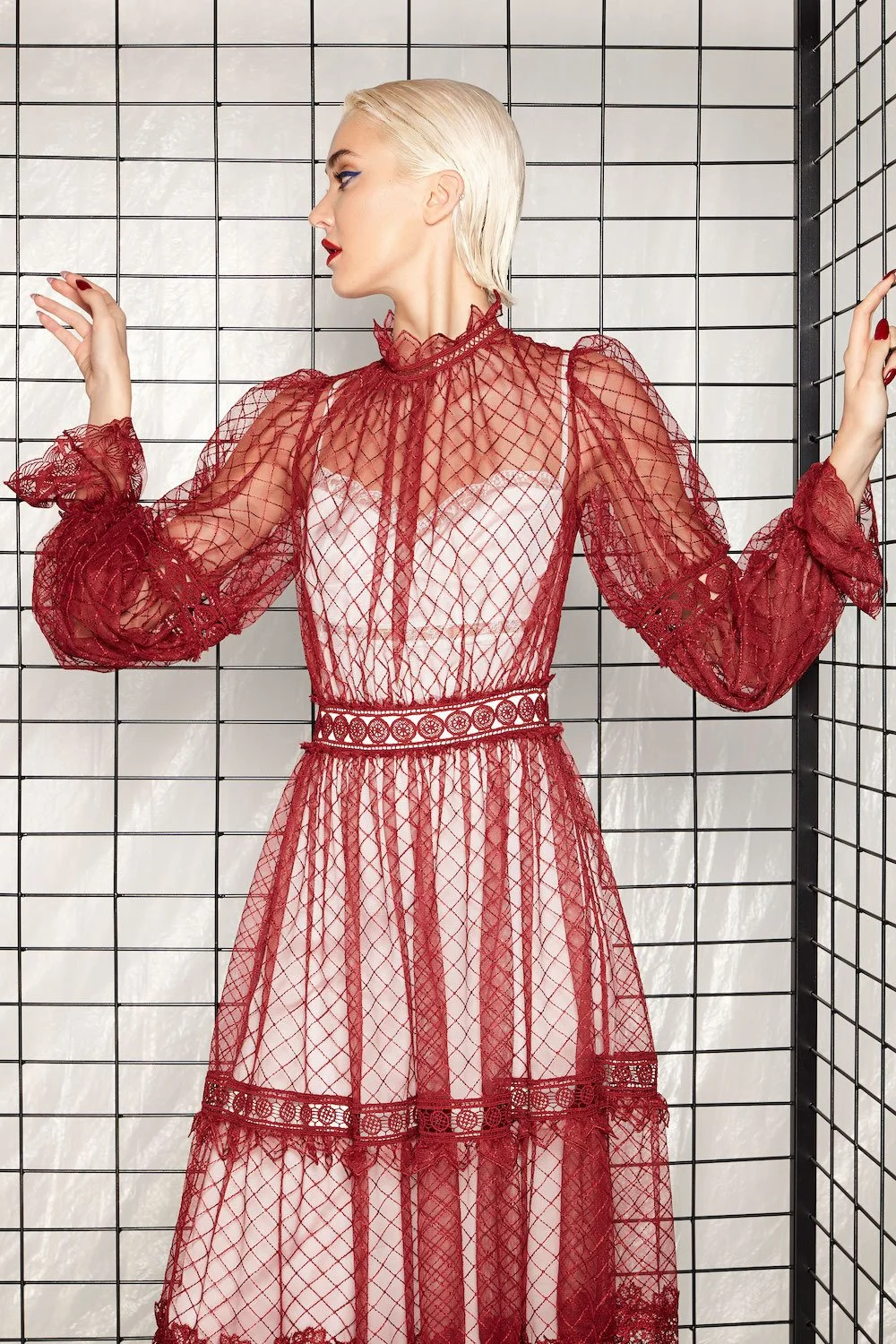 A woman with platinum blonde hair wearing a sheer red lace dress with intricate patterns, standing against a black wire grid backdrop.