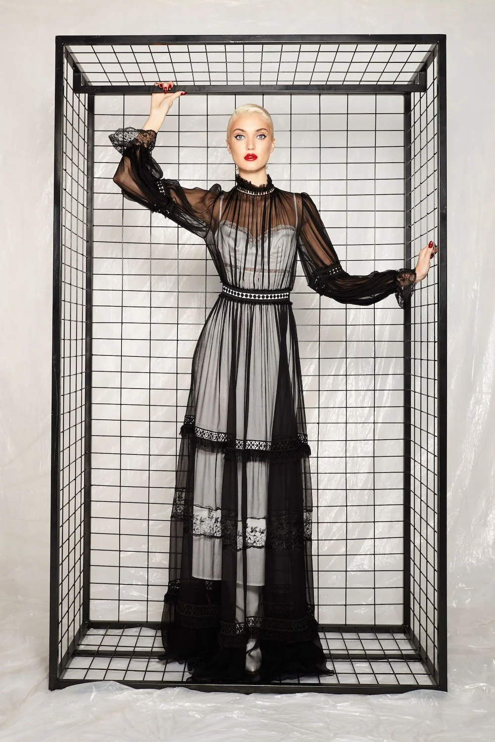 A woman with platinum blonde hair and red lipstick posing inside a black metal grid enclosure, wearing a sheer black and white lace dress with long sleeves and intricate detailing.