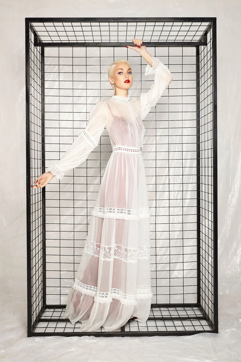 A woman with platinum blonde hair and red lipstick standing inside a black metal grid enclosure, wearing a sheer white dress with lace details.