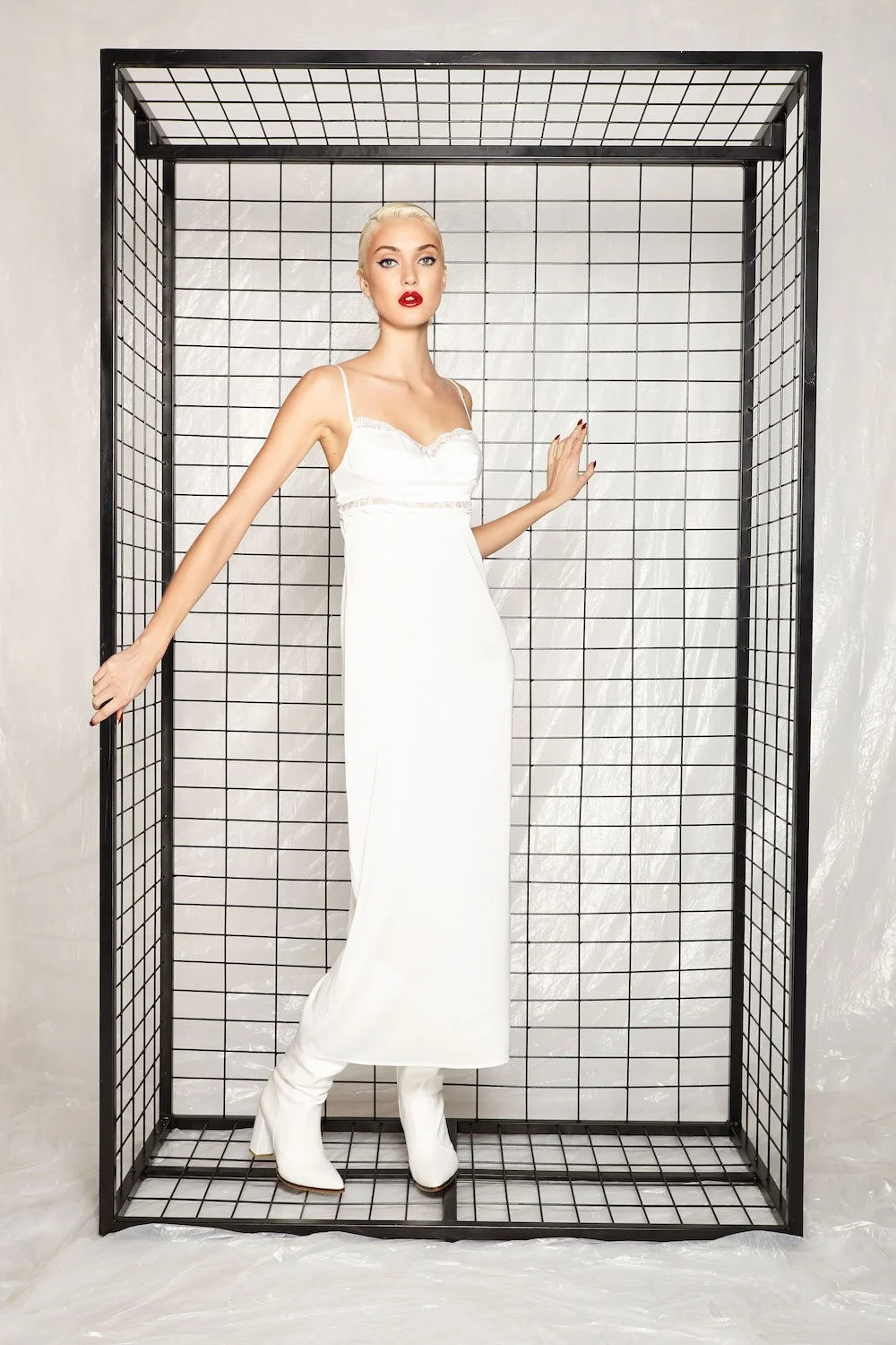 A woman in a white dress and white boots standing inside a black wireframe cube with a marble wall background.