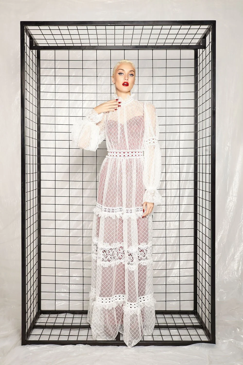 Woman in a sheer white lace dress with long sleeves, standing inside a black metal frame with a grid pattern.