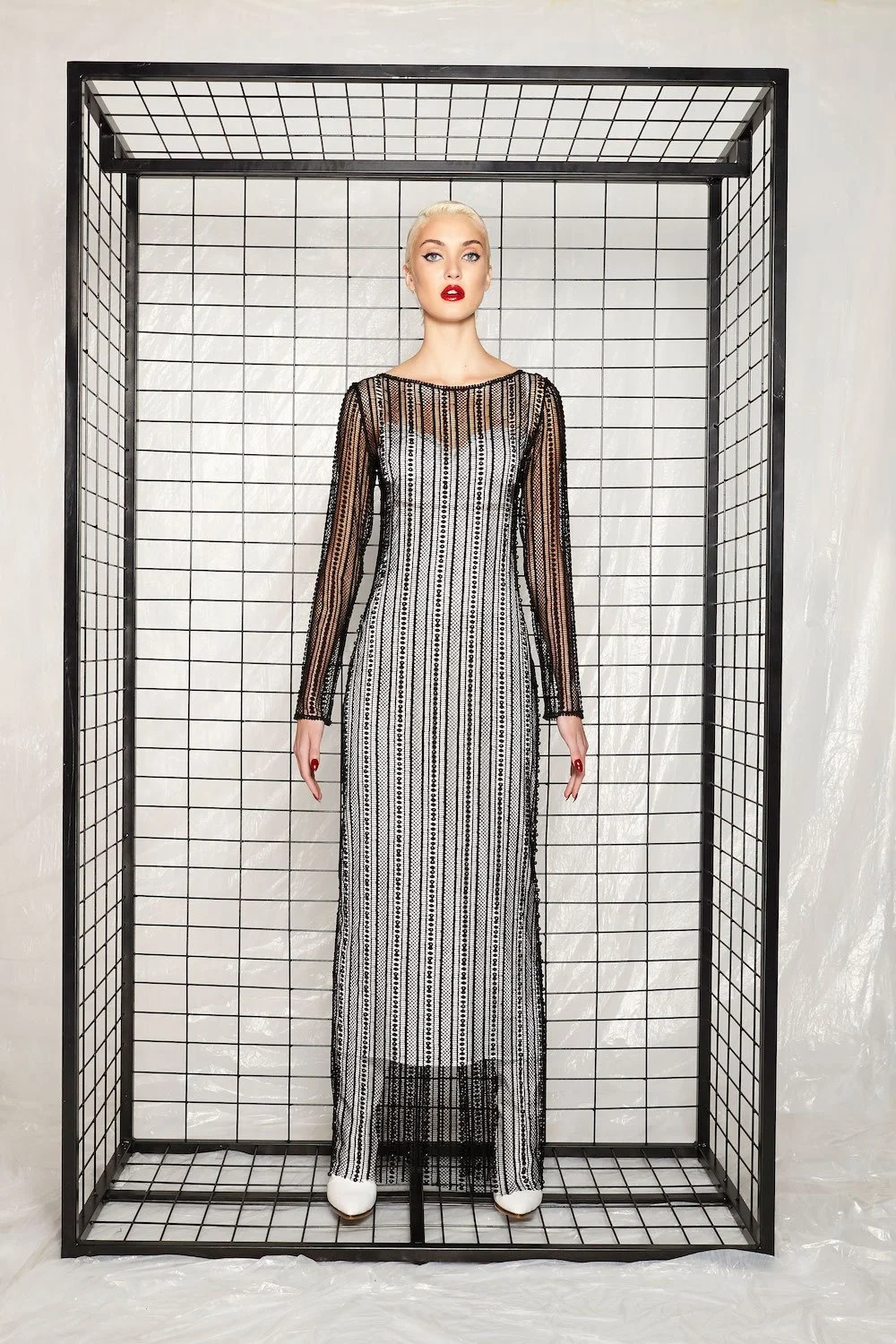A woman with platinum blonde hair, red lipstick, and makeup, wearing a long, black-and-white striped, sheer dress with detailed patterns, stands inside a black wireframe cage against a plain, light background.
