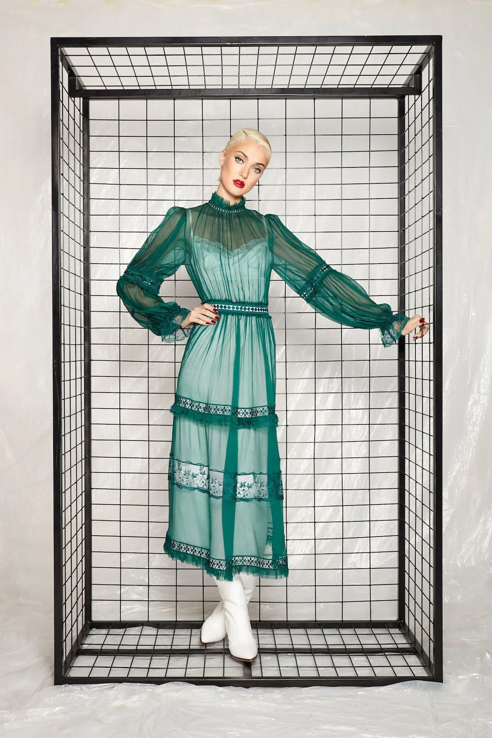 A woman with platinum blonde hair and red lipstick wearing a teal, sheer, long-sleeved, floor-length dress with lace detailing, posing inside a black wireframe rectangular enclosure with a light-colored background.