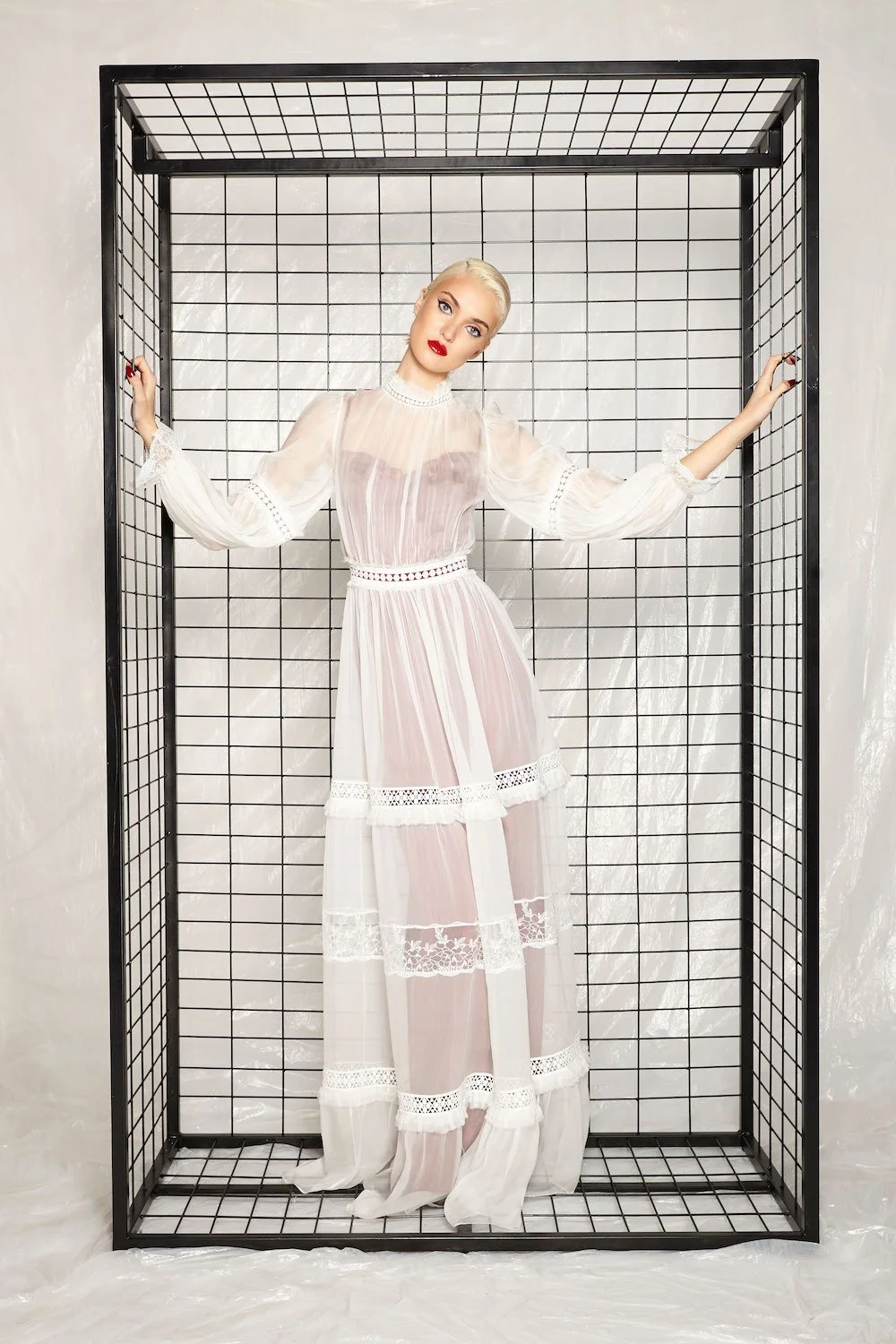 Woman in a sheer white dress standing inside a black metal cage with a grid pattern, posed with arms outstretched.
