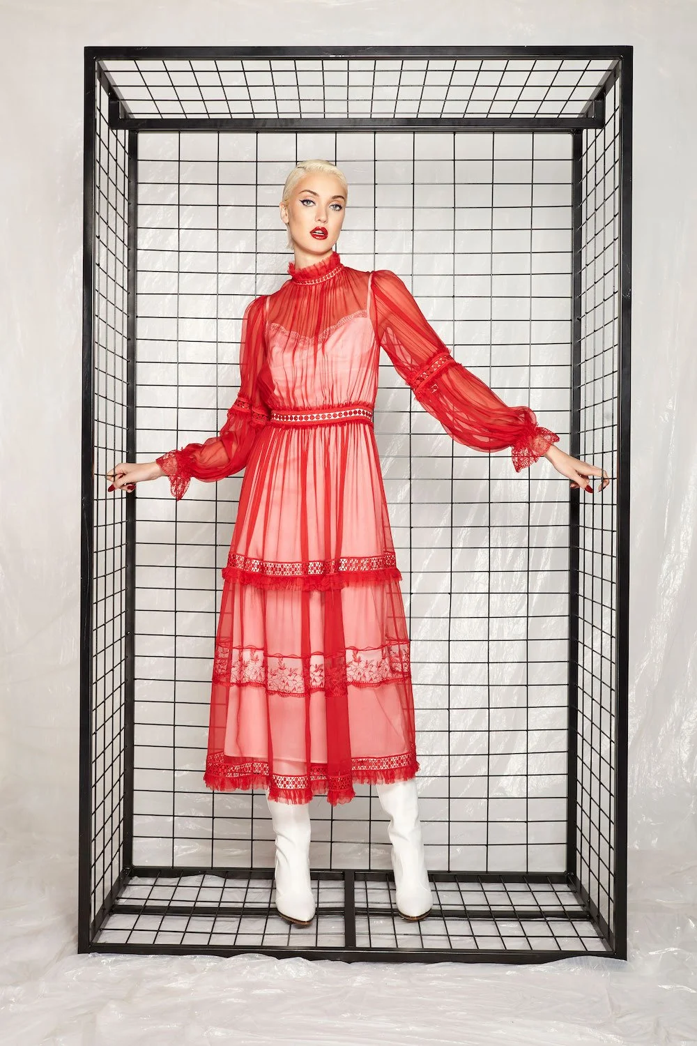 Woman with platinum blonde hair wearing a long, sheer red dress with lace details, standing inside a black wireframe cube.