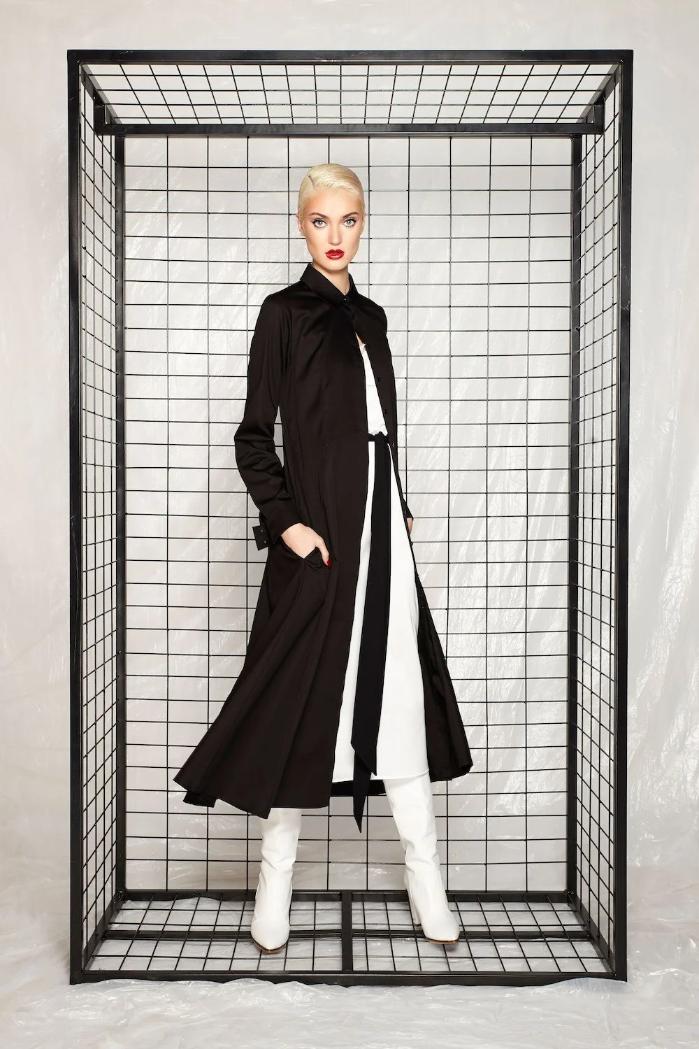 A woman with platinum blonde hair and red lipstick wearing a long black coat, white dress, and white thigh-high boots standing inside a black wireframe enclosure against a white background.