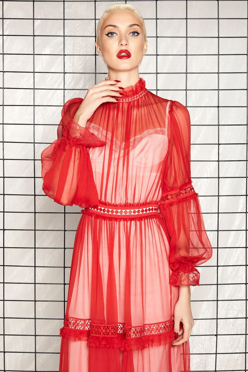 A woman with platinum blonde hair and bold makeup wearing a sheer red dress with lace and ribbon details, standing against a grid-patterned background.