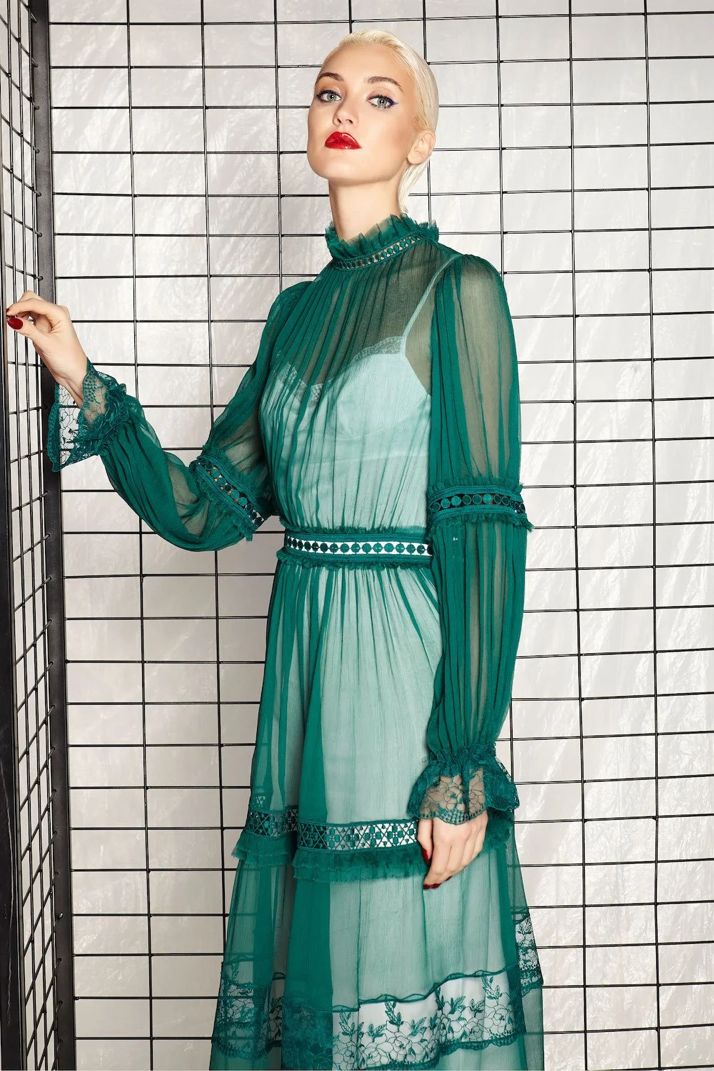 A woman with platinum blonde hair and red lipstick standing in front of a black wire grid, wearing a sheer teal dress with lace and cut-out details.