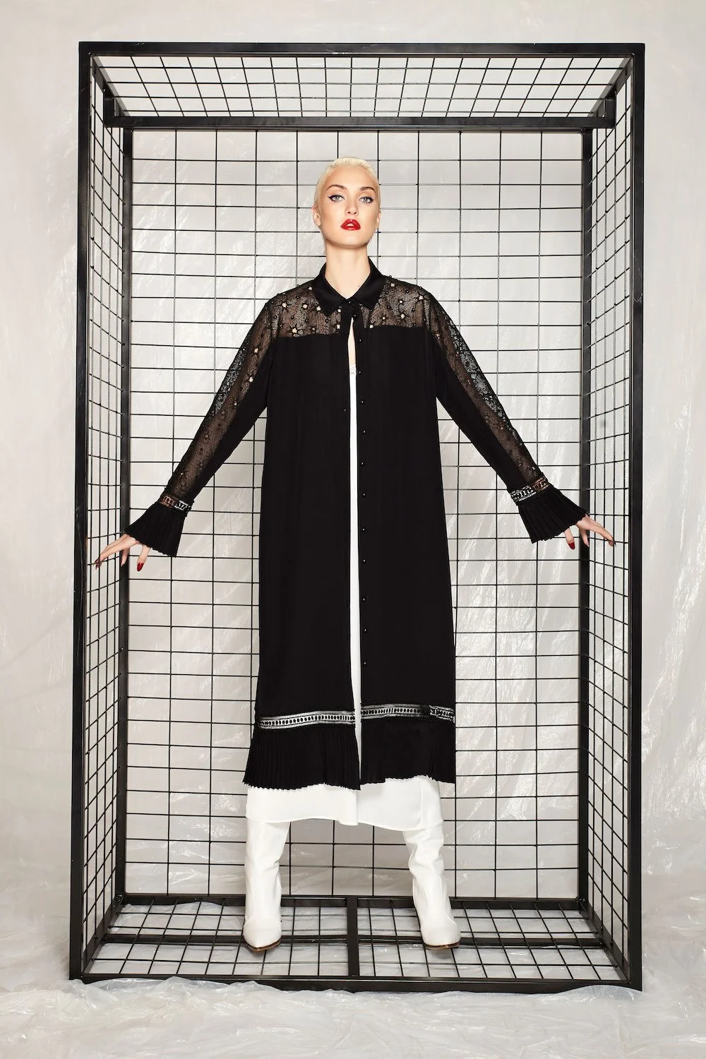 A woman with platinum blonde hair and red lipstick standing inside a black metal grid structure, wearing a long black dress with lace and sheer details on the top and sleeves, and white boots.