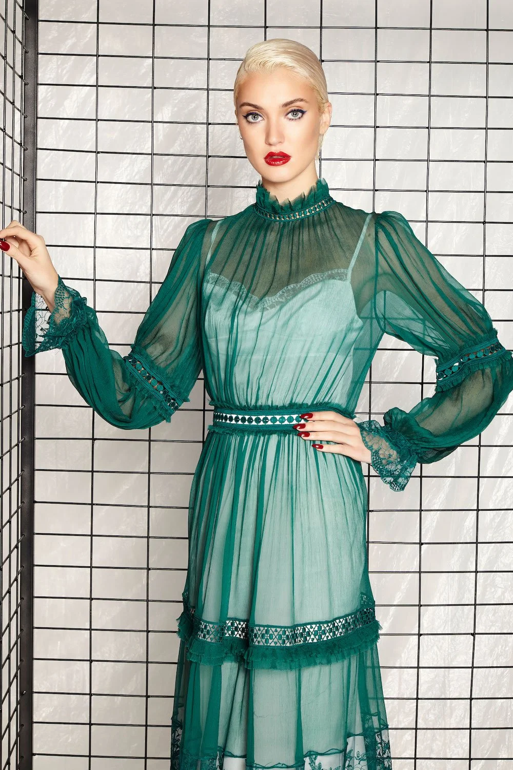 A woman with platinum blonde hair and red lipstick stands against a grid backdrop, wearing an elegant sheer green dress with lace details and ruffled collar.