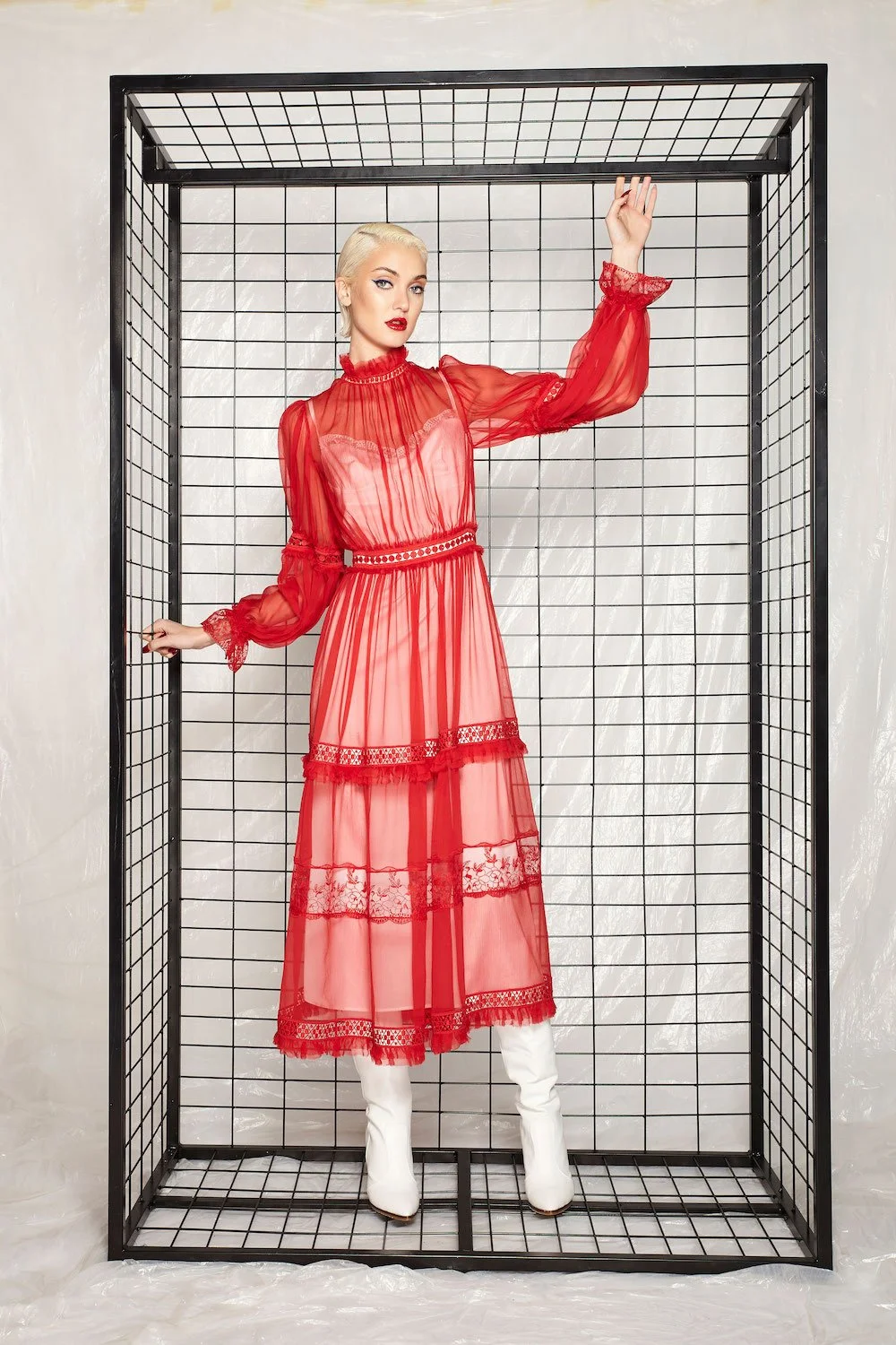 A woman in a sheer red dress with lace details, white boots, and platinum blonde hair, standing within a black wireframe cube structure.