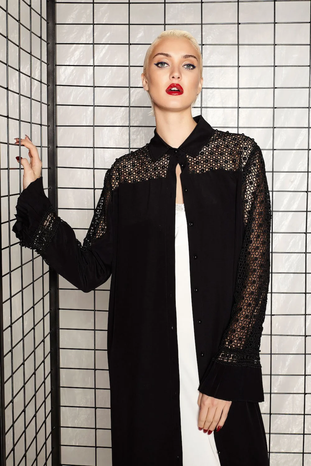 A woman with platinum blonde hair and red lipstick is posing in front of a metal grid backdrop. She is wearing a black shirt with lace detailing on the shoulders and sleeves, and a white garment underneath.