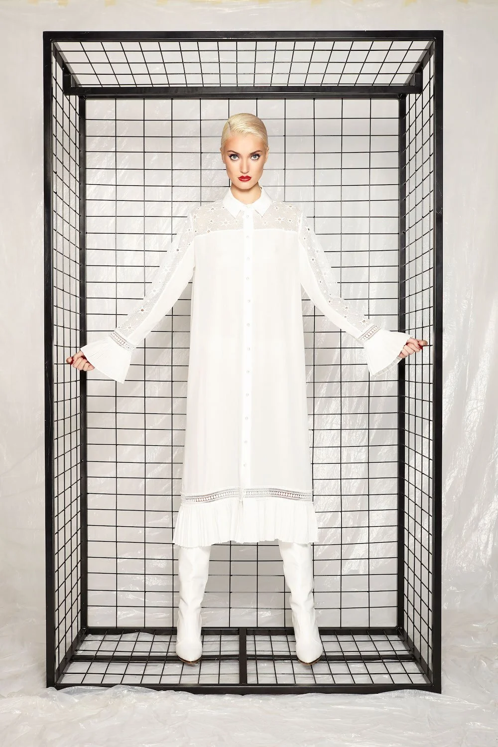 A woman with platinum blonde hair and bold makeup wears a long white dress with sheer lace details and bell sleeves, standing inside a black wireframe enclosure.